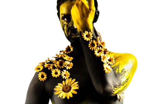 Un body painting très travaillé, avec des fleurs et de la couleur noir et or jaune, avec un arrière plan blanc. Margaux nous observe tout en se dissimulant partiellement le visage.
