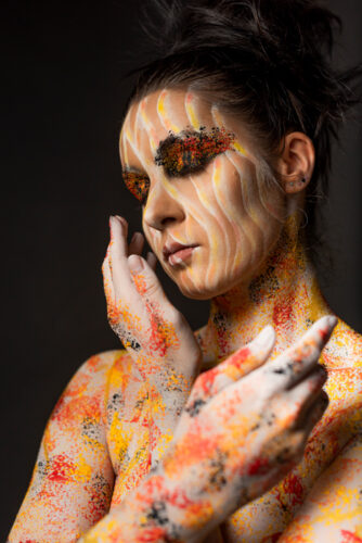 Body painting: la femme aux yeux fermés. Un maquillage orange, jaune or, avec des paillettes sur les yeux. Margaux pose mains croisées vers son buste.