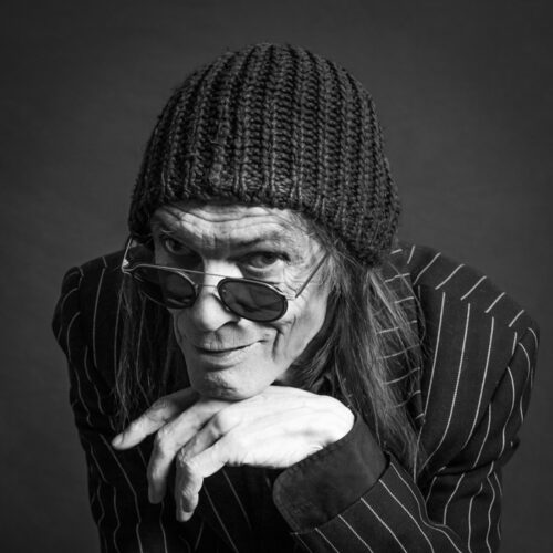 Portrait noir-blanc de Bernie Constantin, chanteur de rock, né en Suisse. Il pose avec la main sous le menton, avec son fidèle bonnet et ses lunettes qui font partie du personnage.