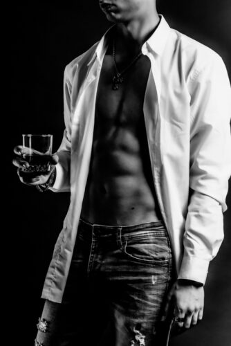 En noir et blanc, le modèle tenant un verre à la main, chemise ouverte sur une musculature mise en exergue par la lumière qui dessine les lignes du corps