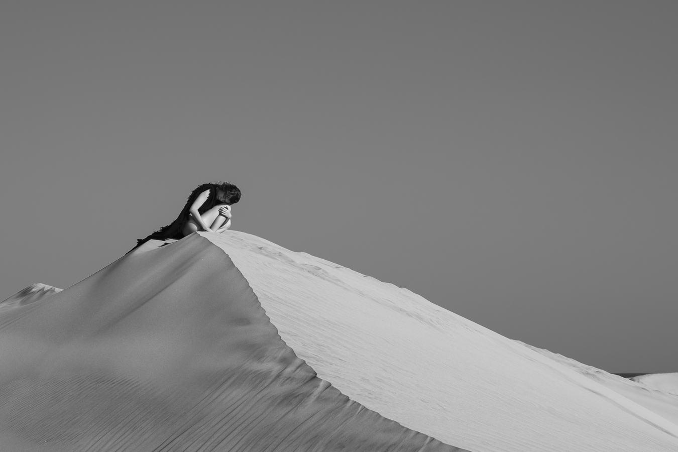 Recroquevillée au sommet de la dune de sable, la modèle semble pensive. Le paysage désertique contraste avec la présence de la femme.