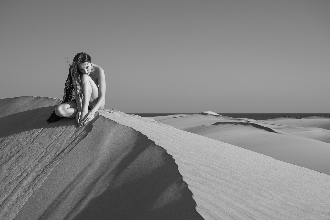 Assise au sommet d'une dune de sable, la modèle pensive, nue, les cheveux dans le vent.
