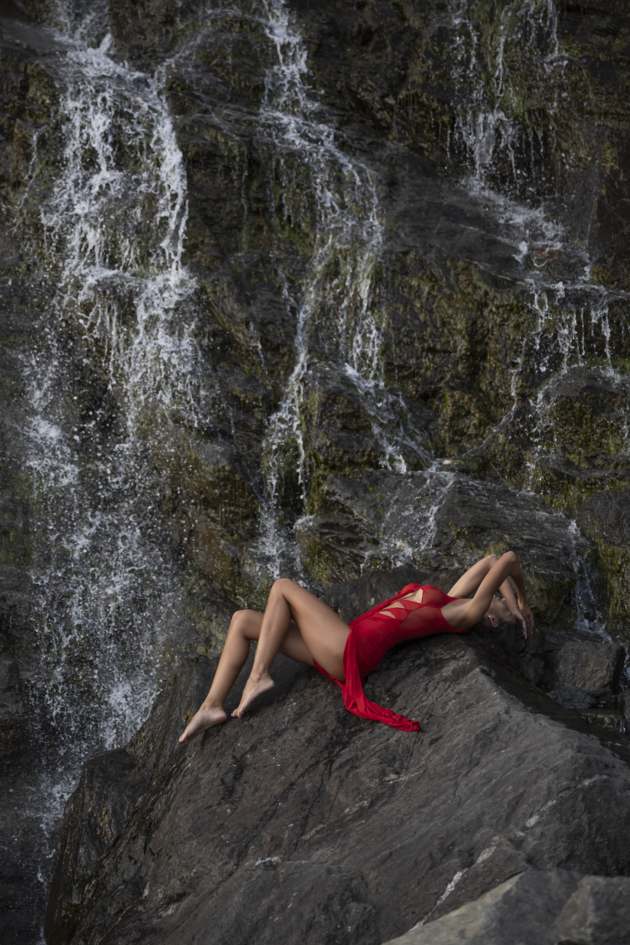 Proche d'une cascade, la modèle est allongée sur un rocher, sa robe rouge contrastant avec la couleur du paysage.