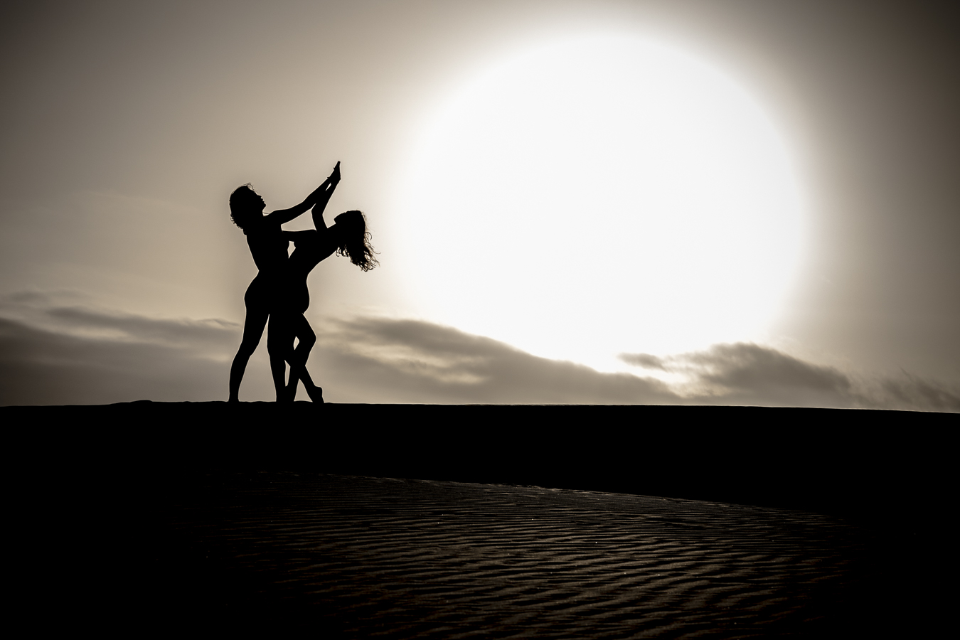 Tezz et Nausicaa Yami enlacées, pour une danse au milieu des dunes de sable. Le soleil couchant, comme une boule de feu dans le ciel, ajoute un peu de mystère à ce lieu